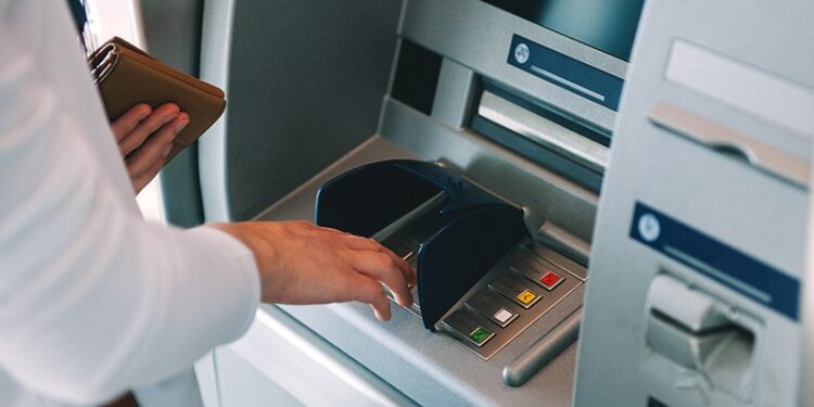 Person using an ATM, inserting a card while holding a wallet nearby in a bank lobby