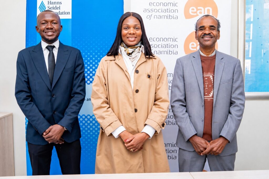 WhatsApp Image 2026-08-02 at 80404 PM - The Brief | Namibia's Leading Business & Financial News Three professionally dressed individuals stand side by side smiling in front of banners for the Economic Association of Namibia (Namibia business event).