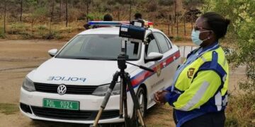 Female police officer in a neon jacket and mask stands beside a white patrol car with a camera on a tripod for speed enforcement.