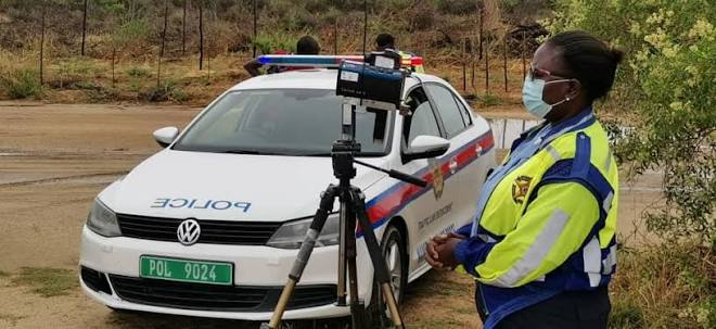 Female police officer in a neon jacket and mask stands beside a white patrol car with a camera on a tripod for speed enforcement.