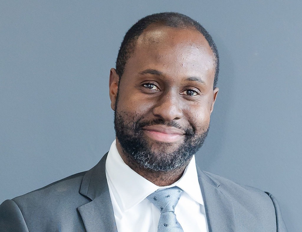 Professional head-and-shoulders portrait of a smiling man in a gray suit and light blue tie, against a muted blue backdrop.