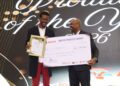 Two men in suits on a stage, smiling as one presents a framed certificate and a large ceremonial check at an awards event with a decorative backdrop.