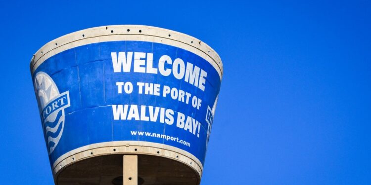 Blue water-tank sign reads 'WELCOME TO THE PORT OF WALVIS BAY' against a clear blue sky, with part of the port logo visible on the left.