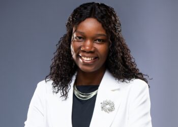 Professional portrait of a smiling Black woman wearing a white blazer, black top, silver necklace, and brooch against a gray backdrop.