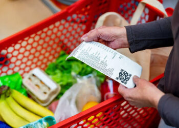 Close-up of a shopper holding a grocery receipt over a red cart filled with bananas, greens, and other groceries.