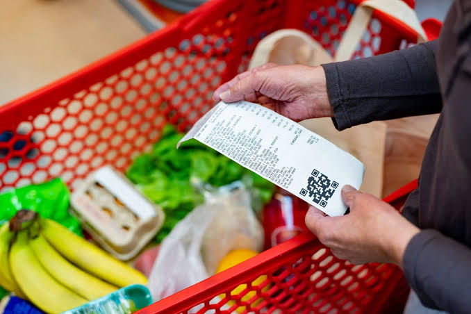 Close-up of a shopper holding a grocery receipt over a red cart filled with bananas, greens, and other groceries.