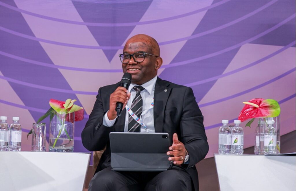 Kashandula satandardbank - The Brief | Namibia's Leading Business & Financial News Man in a dark suit and glasses speaks into a handheld mic while seated on a panel with a tablet on his lap, purple backdrop behind him.
