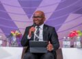 Kashandula satandardbank - The Brief | Namibia's Leading Business & Financial News Man in a dark suit and glasses speaks into a handheld mic while seated on a panel with a tablet on his lap, purple backdrop behind him.