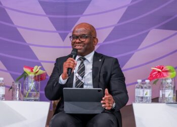 Man in a dark suit and glasses speaks into a handheld mic while seated on a panel with a tablet on his lap, purple backdrop behind him.