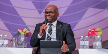 Man in a dark suit and glasses speaks into a handheld mic while seated on a panel with a tablet on his lap, purple backdrop behind him.