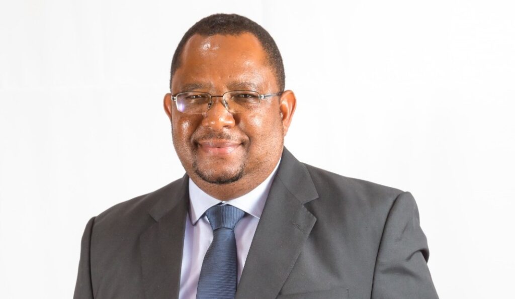 Vehaka Tjimune - The Brief | Namibia's Leading Business & Financial News Professional portrait of a smiling man in a dark suit and blue tie against a white background with glasses on.