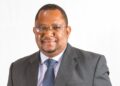 Professional portrait of a smiling man in a dark suit and blue tie against a white background with glasses on.