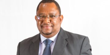 Professional portrait of a smiling man in a dark suit and blue tie against a white background with glasses on.
