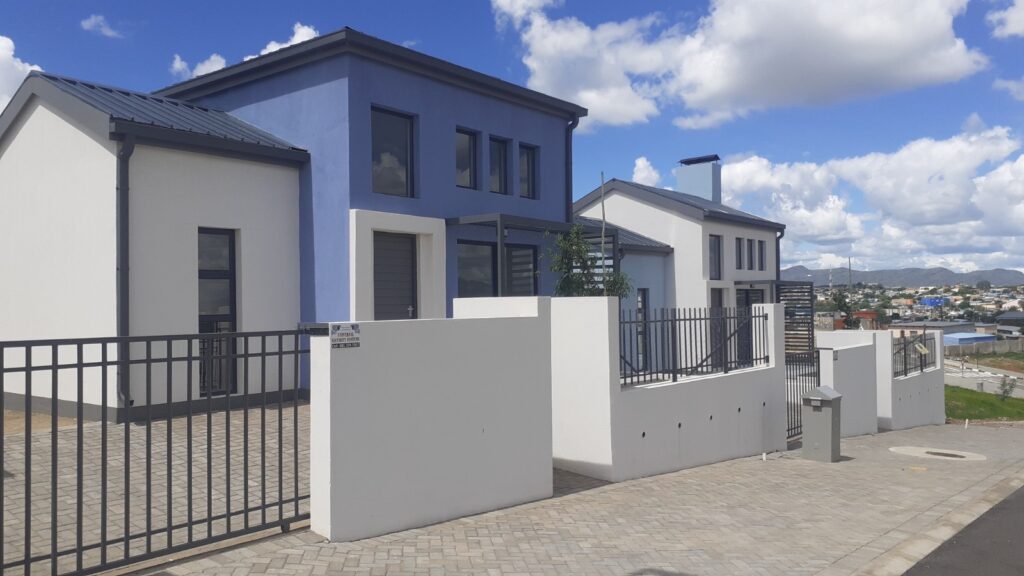 Row of modern white and blue townhouses with metal fences along a paved sidewalk under a bright blue sky with clouds.