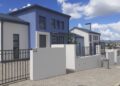 HOUSING5 - The Brief | Namibia's Leading Business & Financial News Row of modern white and blue townhouses with metal fences along a paved sidewalk under a bright blue sky with clouds.