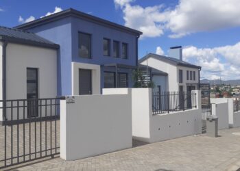 Row of modern white and blue townhouses with metal fences along a paved sidewalk under a bright blue sky with clouds.