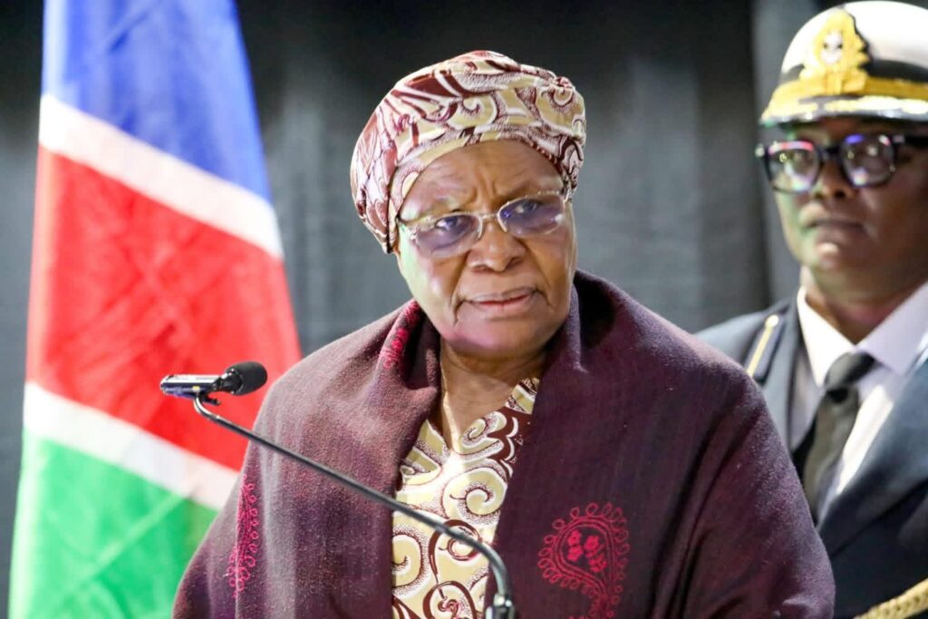 Elderly woman in a patterned headwrap and maroon shawl speaks at a podium with a microphone, flag behind her.