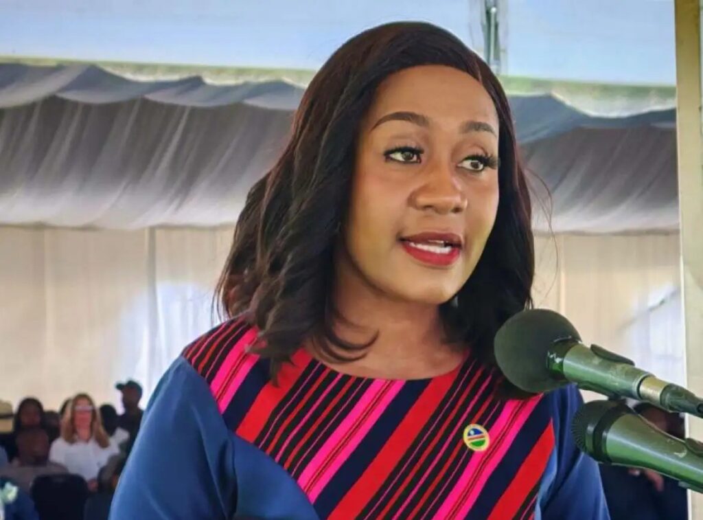 Kadiva Hamutumwa 1 - The Brief | Namibia's Leading Business & Financial News Woman speaking into a microphone at an outdoor event, wearing a blue dress with pink/red diagonal stripes and a badge on her chest.