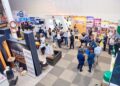 A large trade show hall with groups of people networking at booths along colorful exhibitor banners.