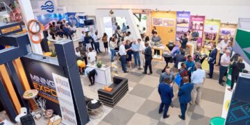 A large trade show hall with groups of people networking at booths along colorful exhibitor banners.