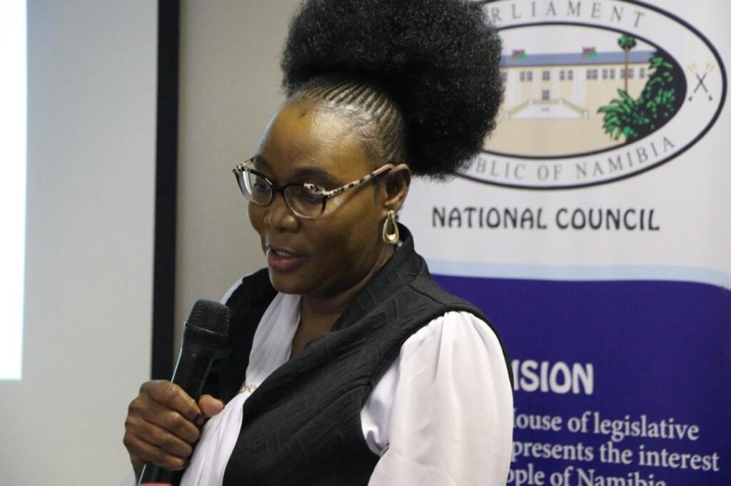 Woman wearing glasses speaks into a handheld microphone at a National Council event, with a banner in the background.