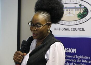 Woman wearing glasses speaks into a handheld microphone at a National Council event, with a banner in the background.