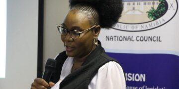 Woman wearing glasses speaks into a handheld microphone at a National Council event, with a banner in the background.