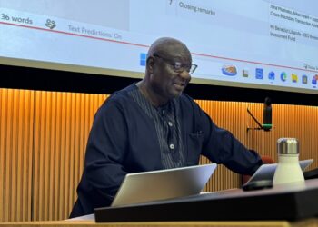 Man in a dark blue traditional outfit presenting at a conference, seated at a desk with a laptop and microphone, a large slide screen behind him.