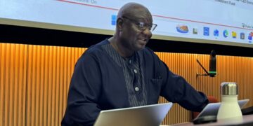 Man in a dark blue traditional outfit presenting at a conference, seated at a desk with a laptop and microphone, a large slide screen behind him.