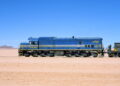 Blue diesel locomotive with yellow stripe parked on a desert track, pulling a railcar under a clear blue sky in a barren landscape.