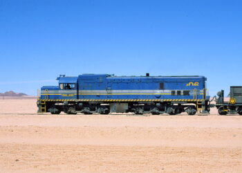 Blue diesel locomotive with yellow stripe parked on a desert track, pulling a railcar under a clear blue sky in a barren landscape.