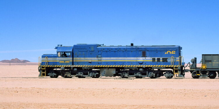 Blue diesel locomotive with yellow stripe parked on a desert track, pulling a railcar under a clear blue sky in a barren landscape.