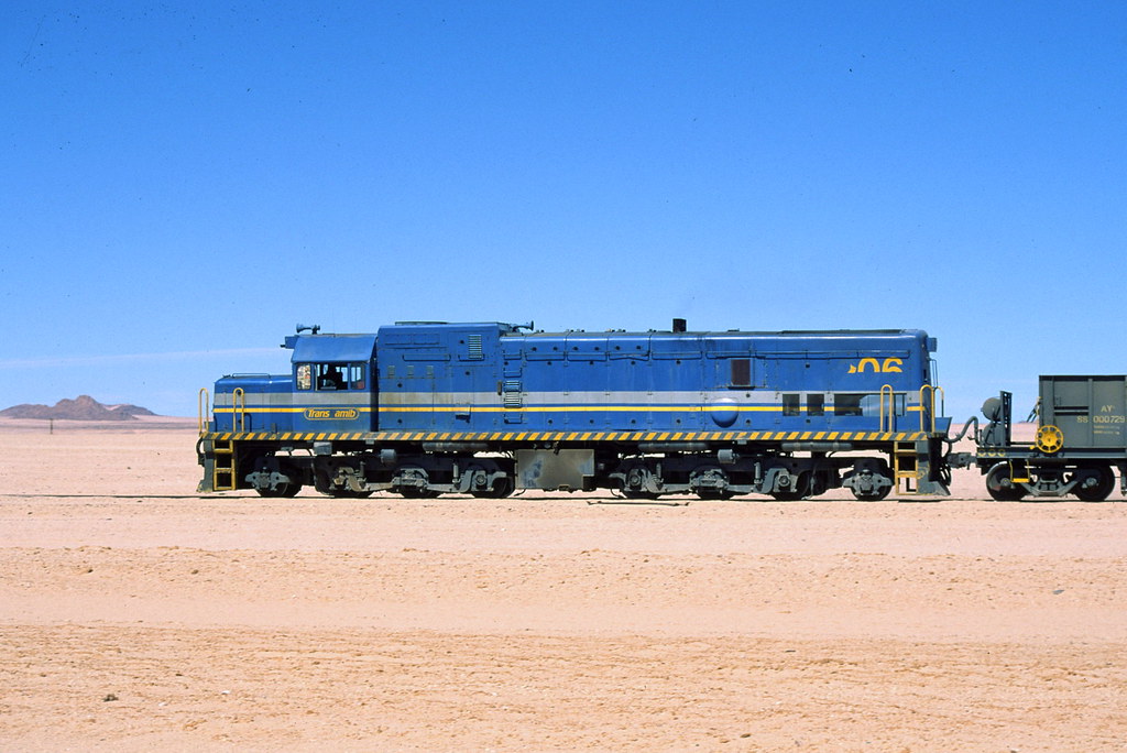 Blue diesel locomotive with yellow stripe parked on a desert track, pulling a railcar under a clear blue sky in a barren landscape.