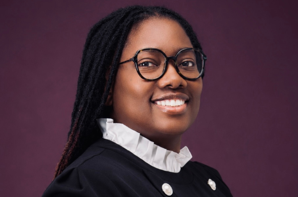 Smiling woman with round glasses and a white ruffled collar against a plum background.