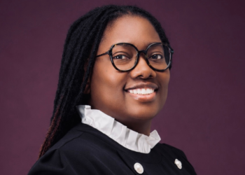 Smiling woman with round glasses and a white ruffled collar against a plum background.