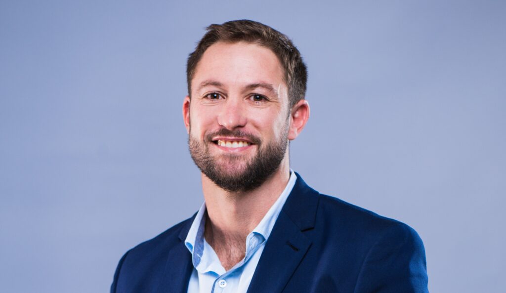 Michael Nel - The Brief | Namibia's Leading Business & Financial News Smiling man in a navy blazer and light blue shirt against a solid blue background (portrait).
