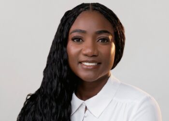 Monika Amukoto - The Brief | Namibia's Leading Business & Financial News Portrait of a smiling Black woman with long braided hair wearing a white collared shirt against a light gray background, studio portrait