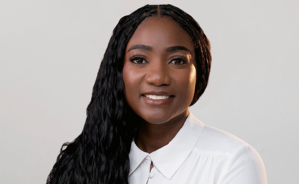 Monika Amukoto - The Brief | Namibia's Leading Business & Financial News Portrait of a smiling Black woman with long braided hair wearing a white collared shirt against a light gray background, studio portrait