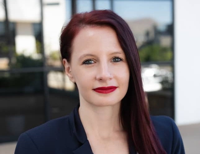 Portrait of a woman with burgundy hair and bright red lipstick, wearing a navy blazer outdoors.