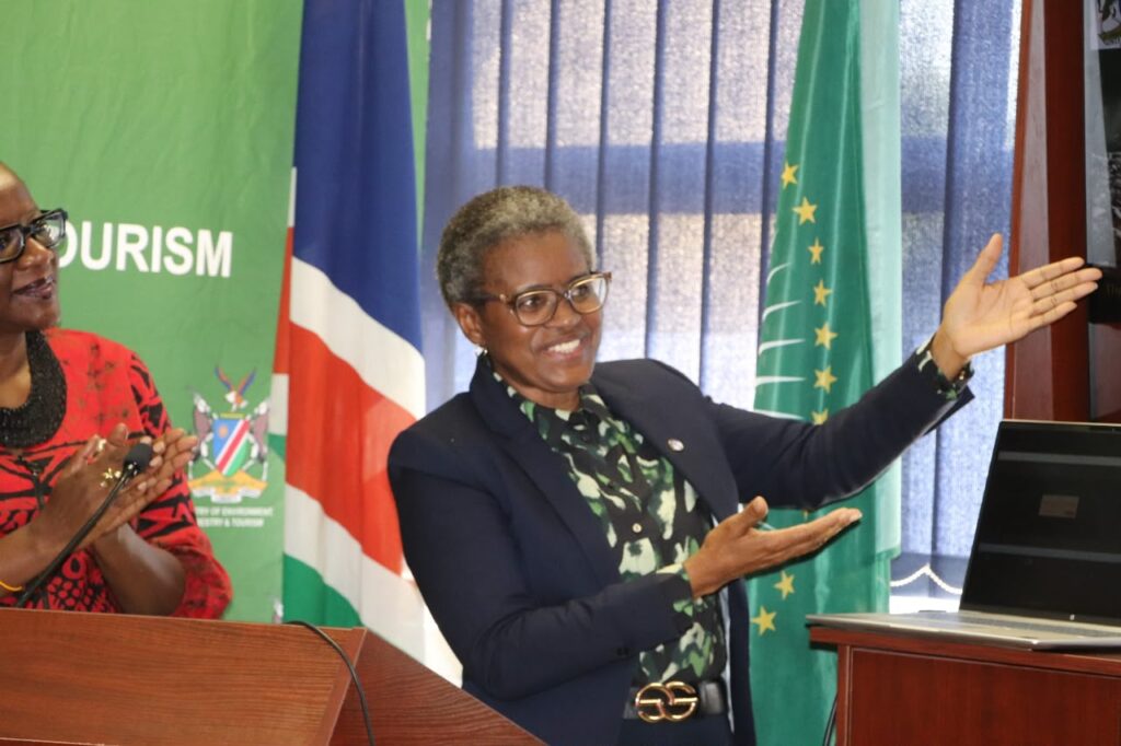 Two women at a conference: one in a navy blazer gesturing toward a laptop, with flags and a green tourism banner in the background.
