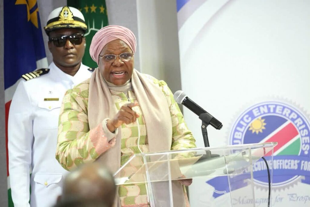 Woman in a pink headscarf and pastel suit speaking at a podium with a microphone; a uniformed officer stands behind her.