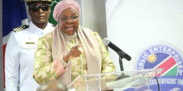 Woman in a pink headscarf and pastel suit speaking at a podium with a microphone; a uniformed officer stands behind her.