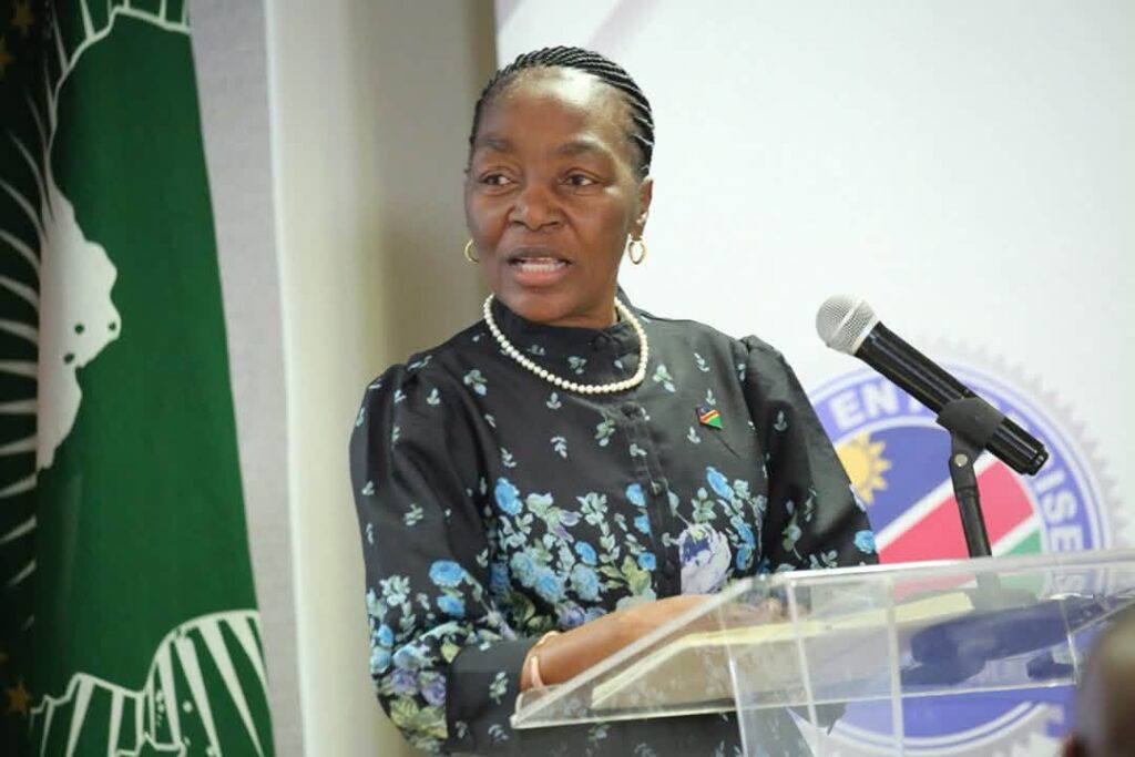 ERICAH9 - The Brief | Namibia's Leading Business & Financial News A woman in a floral dark dress with a pearl necklace speaks at a transparent podium with a microphone, a flag pin on her chest.