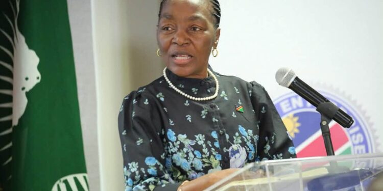 A woman in a floral dark dress with a pearl necklace speaks at a transparent podium with a microphone, a flag pin on her chest.