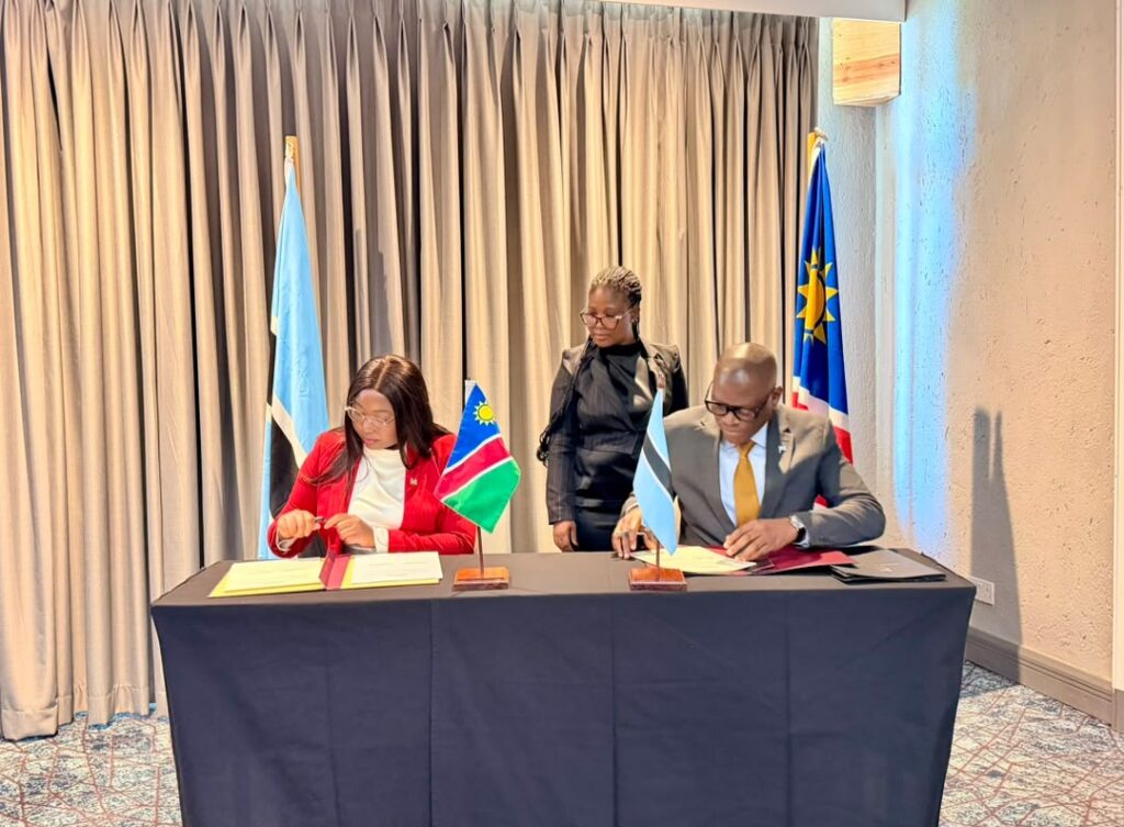 Two officials sign documents at a table with small flags (Namibia and Botswana) as a colleague looks on in a formal signing ceremony behind tall curtains and national flags.