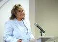 Woman in a light blue blazer speaks at a clear acrylic podium with a microphone, smiling as she presents.