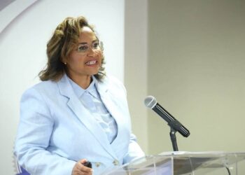 Patty Karuaihe-Martin2 - The Brief | Namibia's Leading Business & Financial News Woman in a light blue blazer speaks at a clear acrylic podium with a microphone, smiling as she presents.