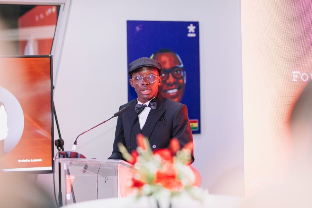 Man in a black suit and bow tie giving a formal speech at a podium with a microphone, wearing a flat cap and round glasses.
