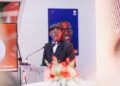 KC Picture - The Brief | Namibia's Leading Business & Financial News Man in a black suit and bow tie giving a formal speech at a podium with a microphone, wearing a flat cap and round glasses.