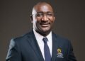 Professional head-and-shoulders portrait of a smiling man in a navy suit and tie, wearing glasses, with a Namibia University of Science and Technology logo on his blazer.
